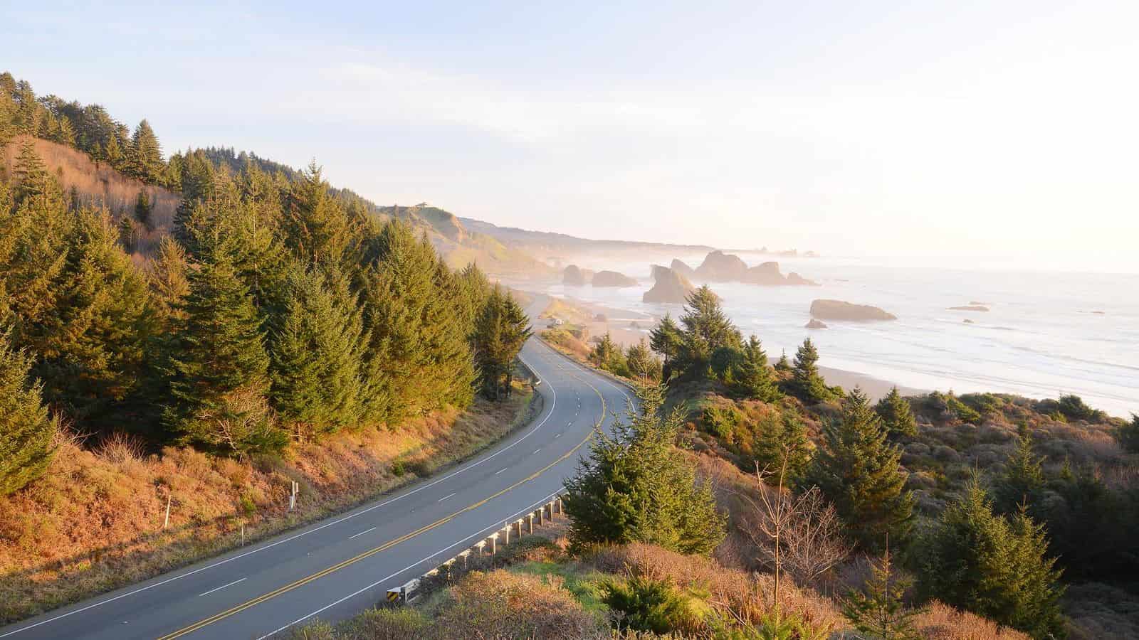 A scenic coastal road, perfect for Thanksgiving road trips, curves through a hilly landscape with dense evergreen trees. The road runs alongside a sandy beach with large rock formations in the distance, under a clear, sunlit sky.