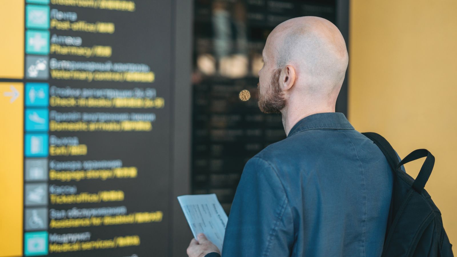 A photo of a man holding passport and looking at boarding screen.