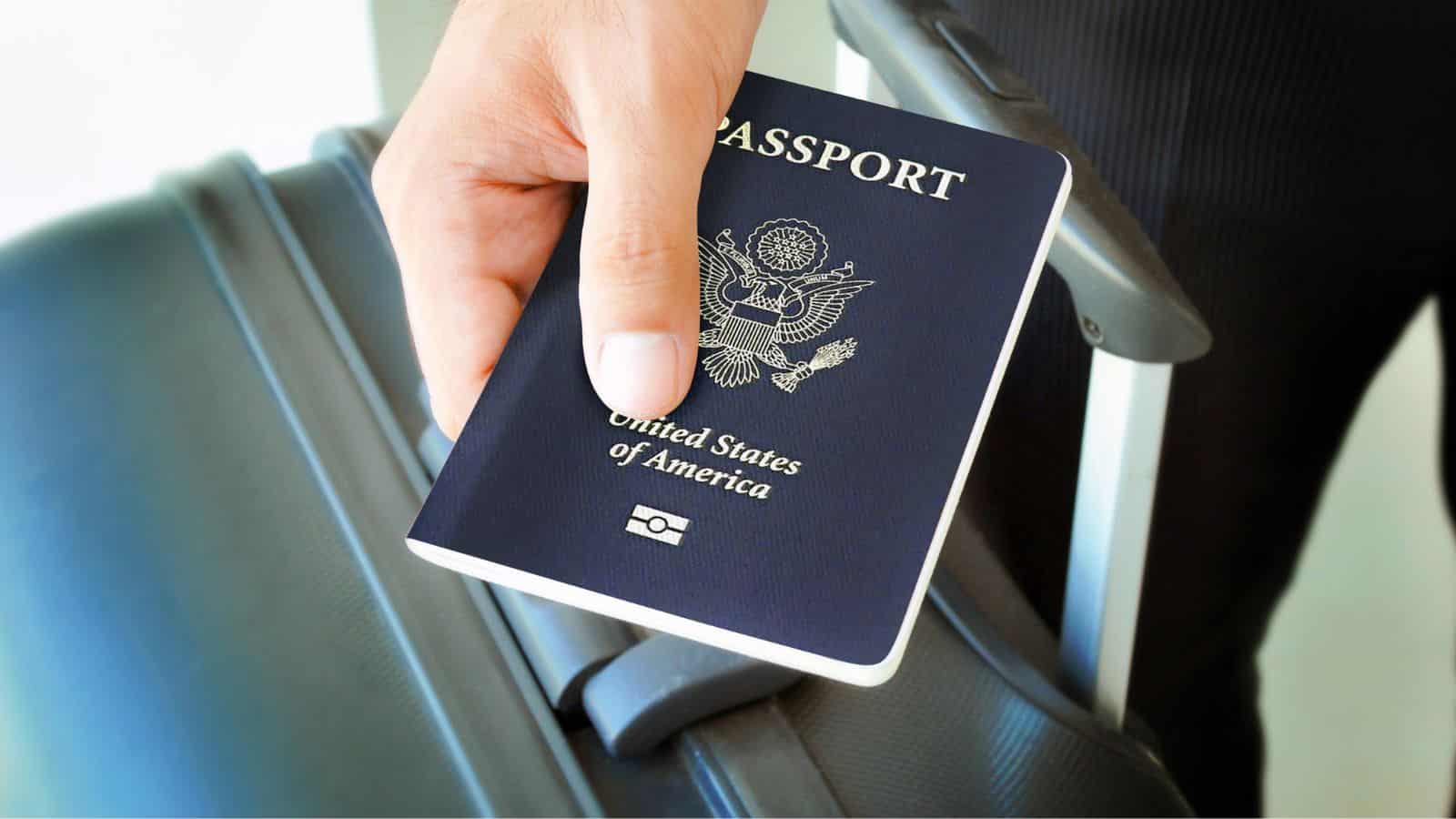 A person holds a U.S. passport and rests their hand on a gray suitcase handle.