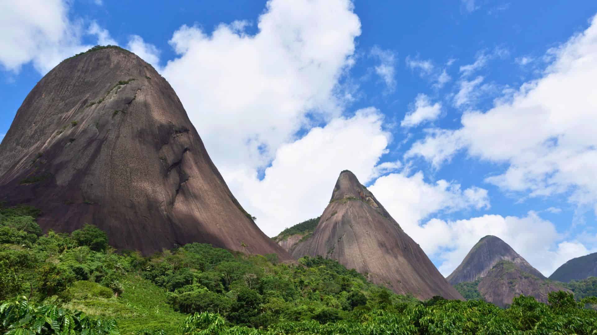 Rocky mountain peaks tower over dense greenery beneath a blue sky dotted with white clouds.