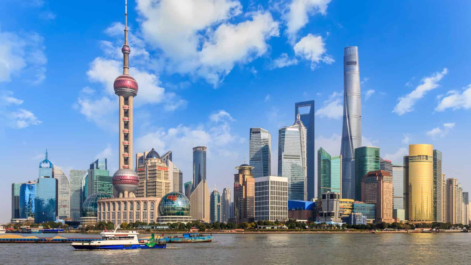 Shanghai skyline featuring modern skyscrapers and the Oriental Pearl Tower beside the Huangpu River under a partly cloudy blue sky.