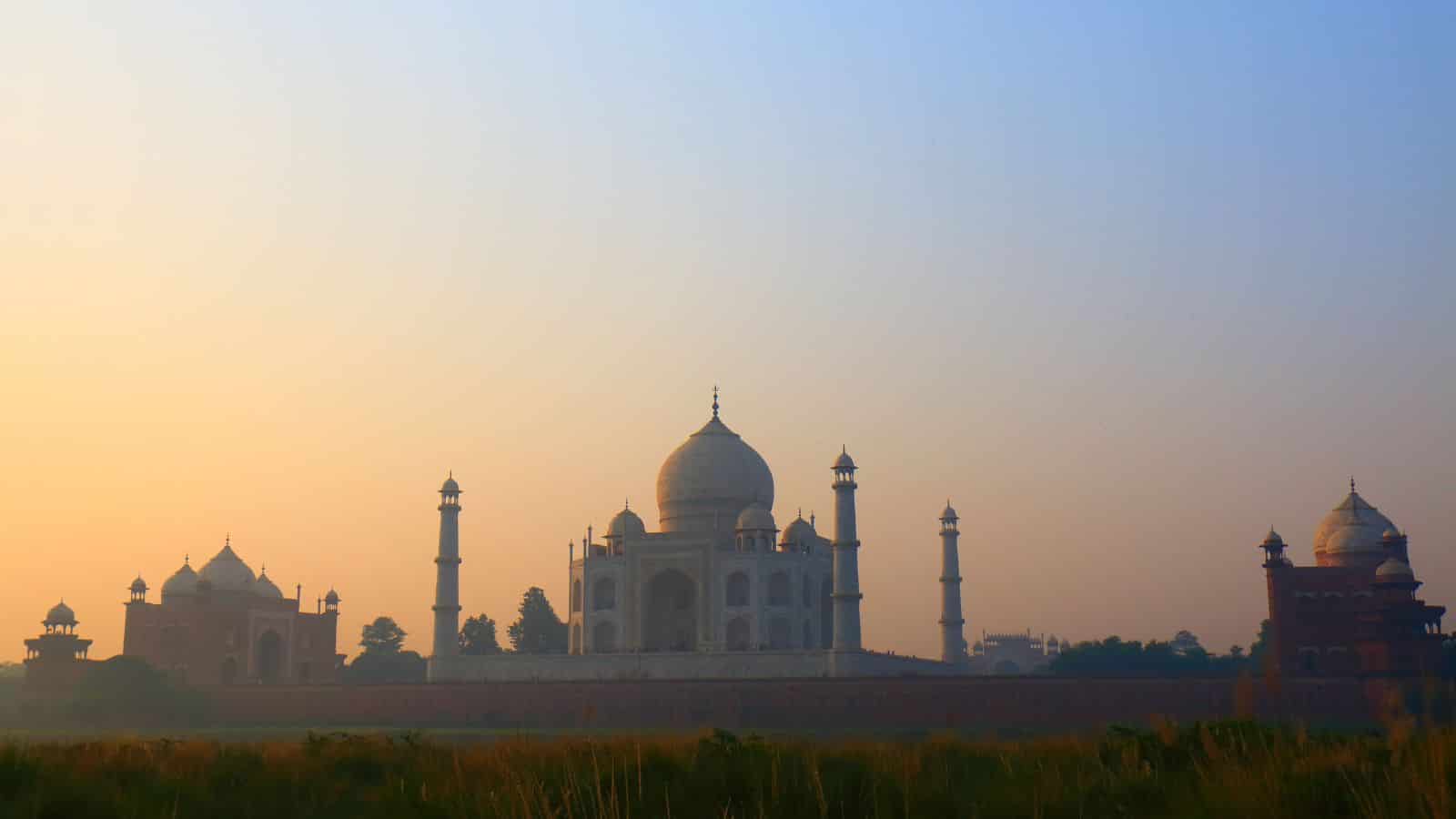 The Taj Mahal and nearby buildings appear as silhouettes before a pastel sky, with grass visible in the foreground.