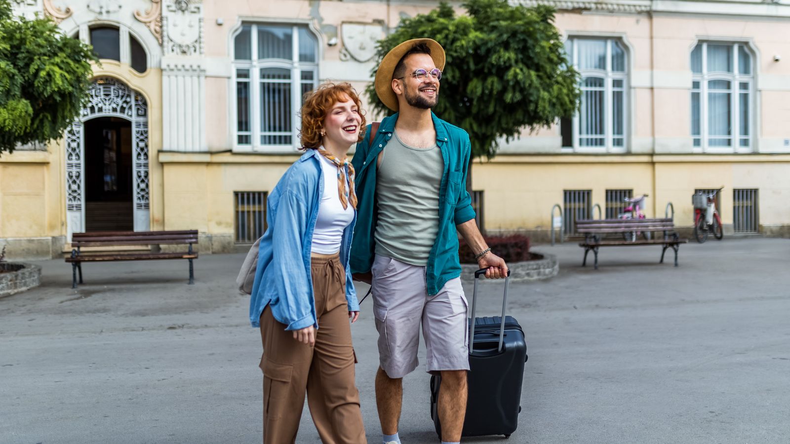 A photo of Young couple traveling together for the first time, walking side by side in a new city, light luggage, soft smiles, warm natural light.