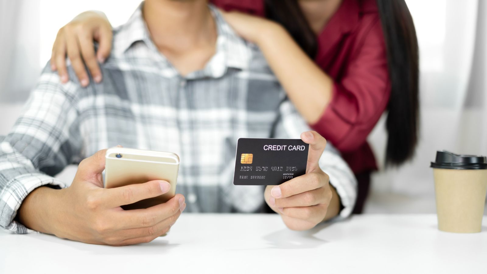 A person holding a smartphone and credit card sits at a table with coffee, while another stands behind with arms on their shoulders.