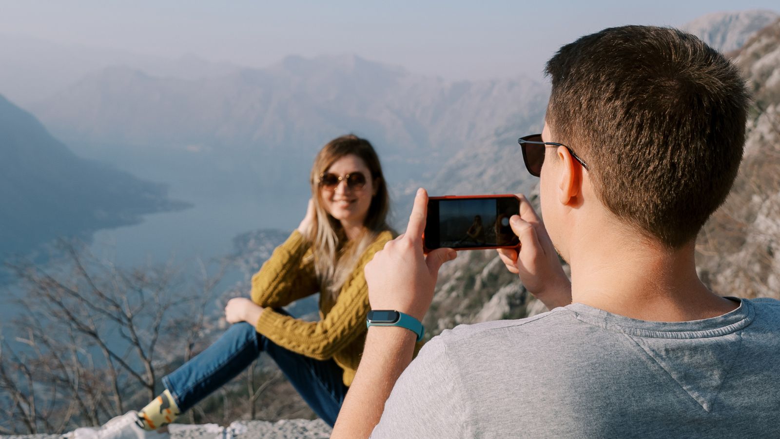 A man photographs a woman seated on a rocky ledge with a lake and mountains in the background on a clear day.