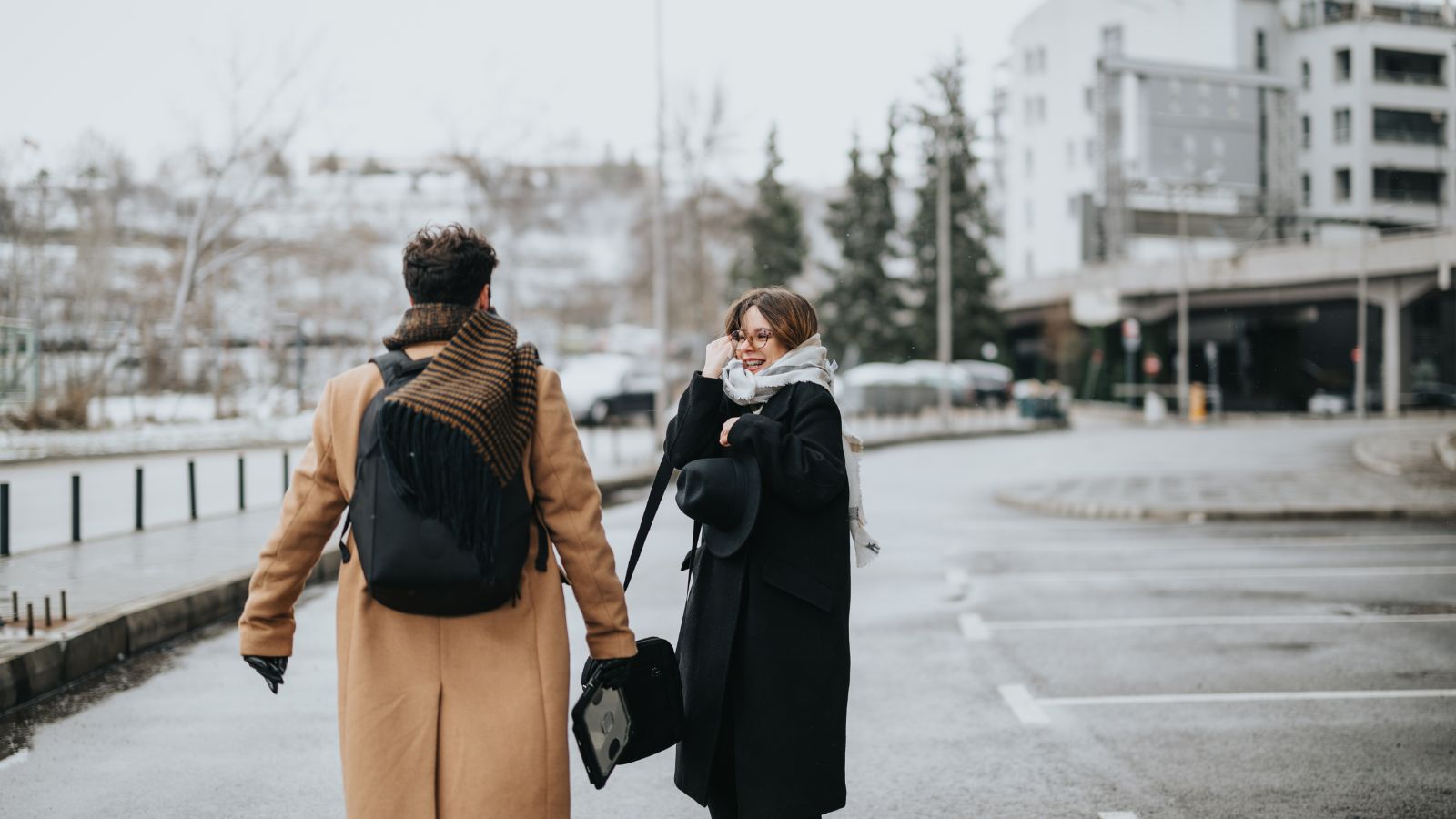 Two people in winter coats stand on a wet city street; one faces away with a bag, the other holds a hat and looks at the camera.