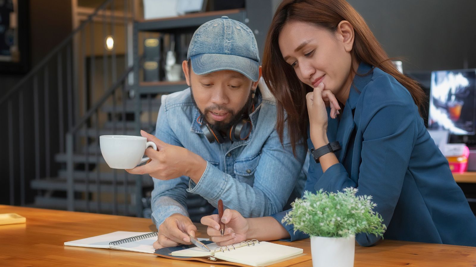Two people sit at a table, one holding a coffee cup as they review notes in a notebook, with a small potted plant nearby.