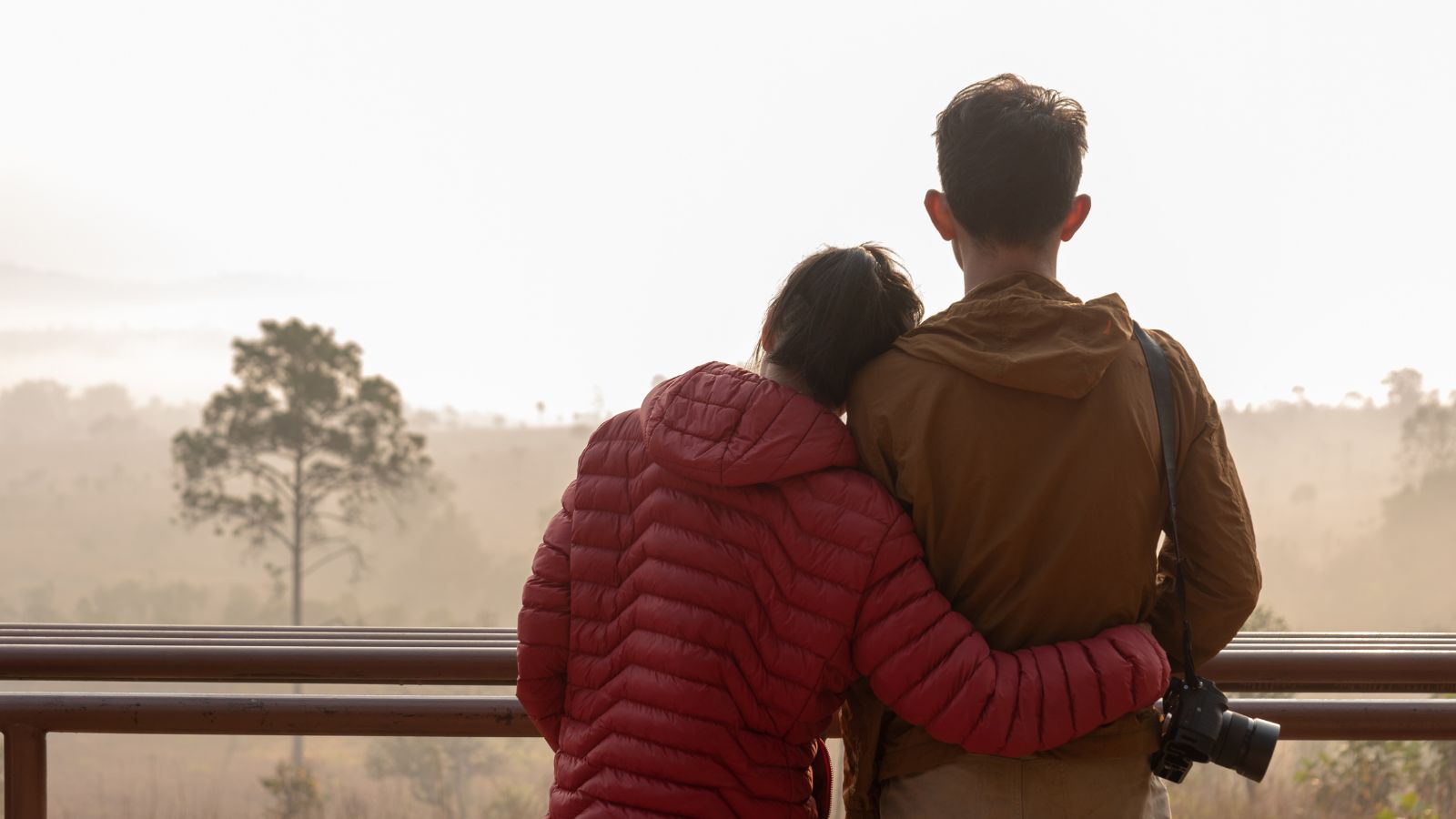 On a foggy balcony, two people in jackets stand close as one leans on the other's shoulder holding a camera.