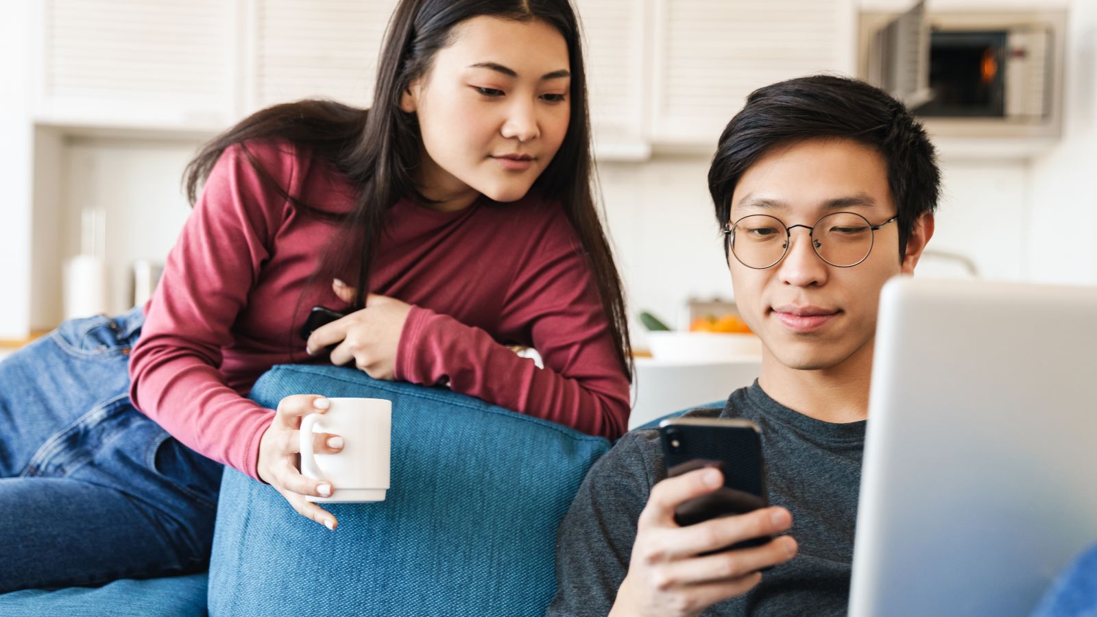 A woman with a mug leans over a couch, watching a man using both a smartphone and laptop at home.