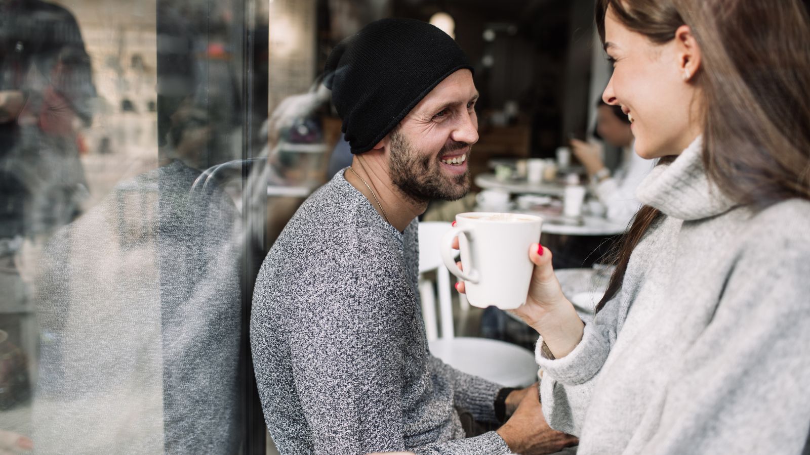 A beanie-wearing man and a woman in a sweater sit together at a café, smiling with coffee mugs in hand.