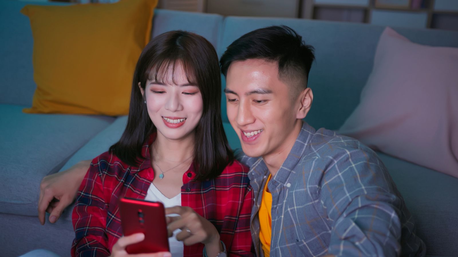 Two people sit on the floor by a couch, smiling at a red smartphone they are holding together.