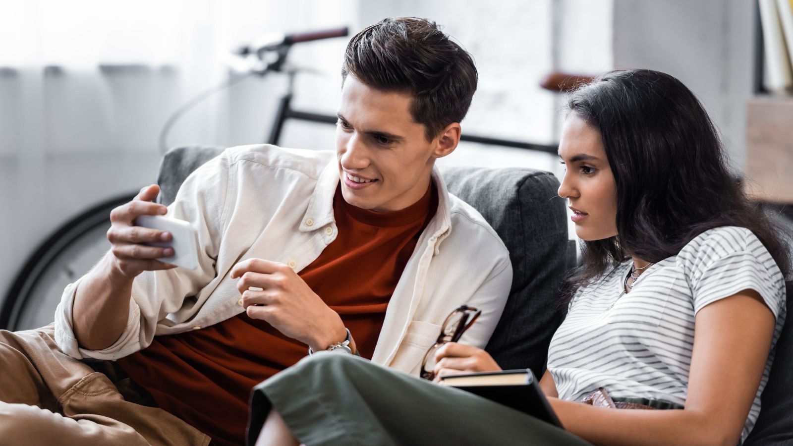 Two people sit on a couch looking at a smartphone; one holds the phone, the other a notebook and glasses, with a bicycle behind them.