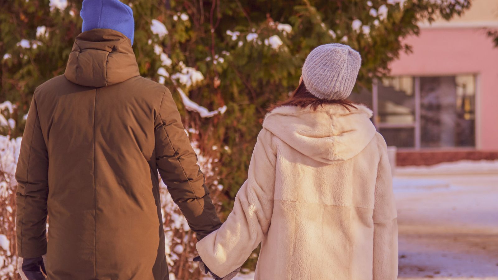 Two people in winter coats and hats walk hand in hand outside on a snowy day, viewed from behind.