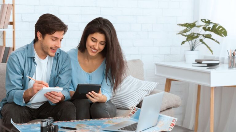 A man and woman sit on a couch viewing a tablet and notebook, with a map and laptop on the table in front of them.