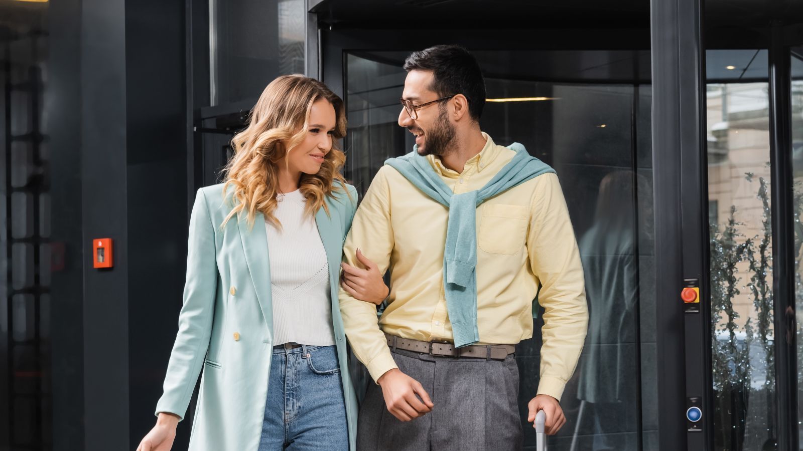 A man and woman walk arm in arm, smiling by a revolving glass door; he has a suitcase, she wears a light blue blazer and jeans.