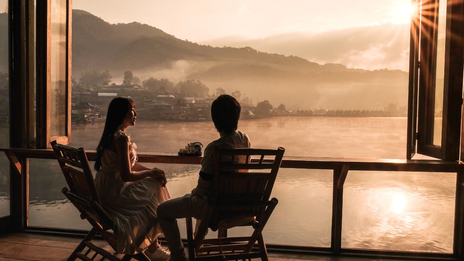 Two people sit on wooden chairs by a lakeside window at sunset, mountains and water visible through the glass.