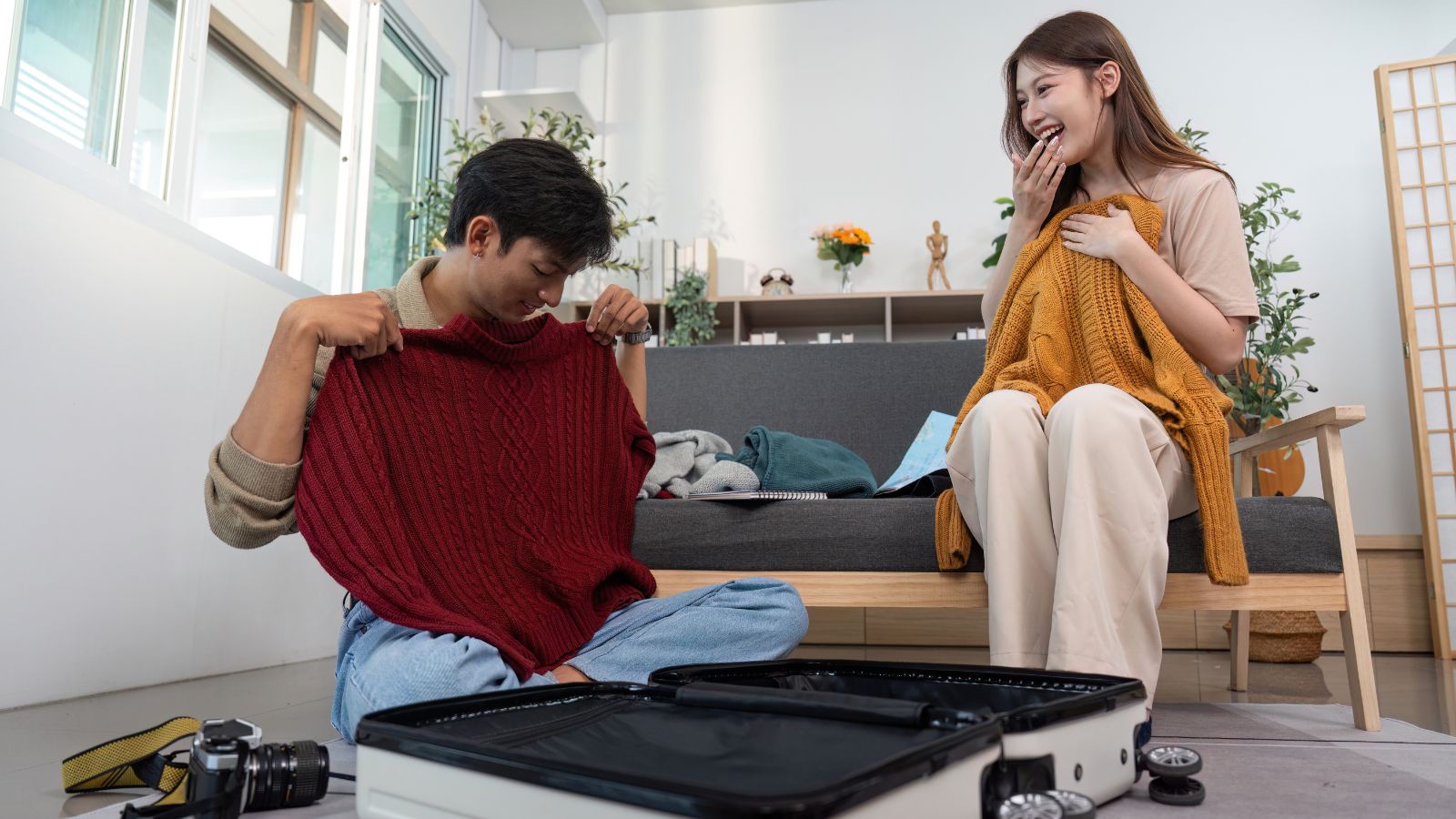 A man and woman pack sweaters into a suitcase on the living room floor, with a camera and open luggage beside them.