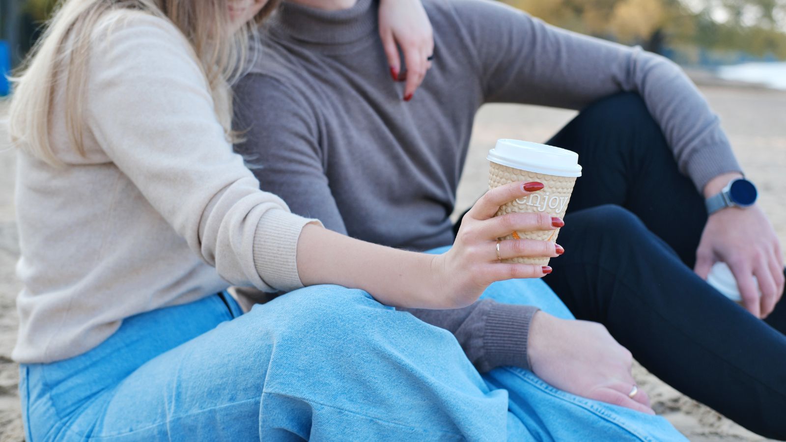 Two people in sweaters and jeans sit close together on sand, with one holding a coffee cup in the foreground.