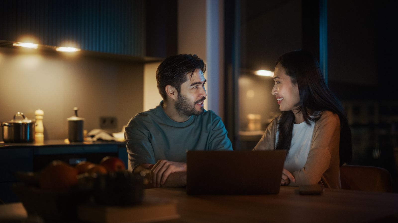 A man and woman sit at a kitchen table with a laptop, talking and smiling in a dimly lit room.