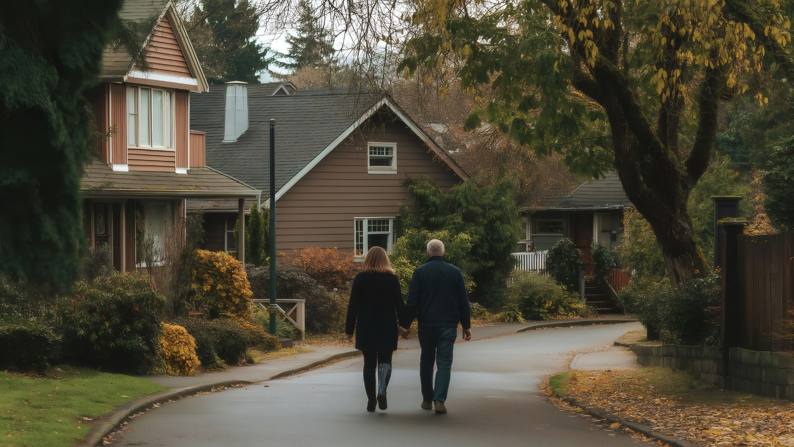 An older couple walks hand in hand along a quiet, tree-lined street lined with houses and autumn leaves.
