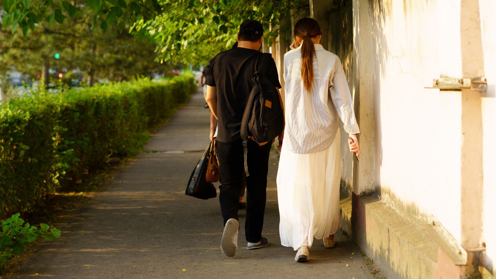 Two people with bags walk away from the camera on a sunlit sidewalk bordered by a wall and greenery.