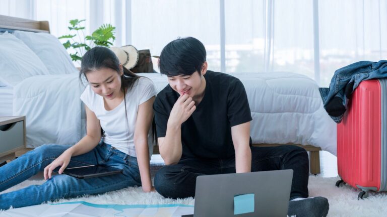Two people sit on a bedroom floor with a laptop, surrounded by papers, a tablet, a red suitcase, and clothes on a chair.