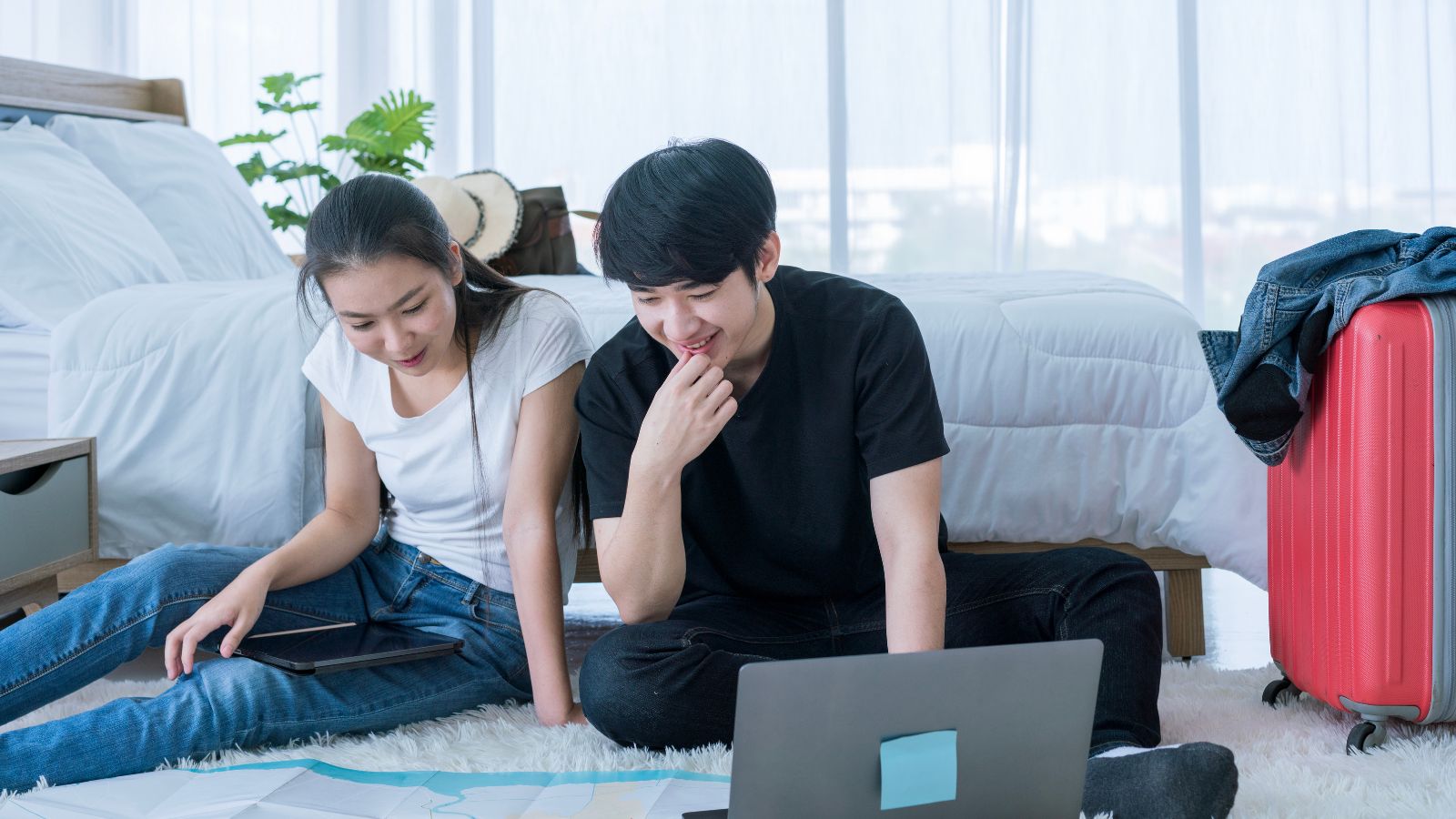 Two people sit on a bedroom floor with a laptop, surrounded by papers, a tablet, a red suitcase, and clothes on a chair.