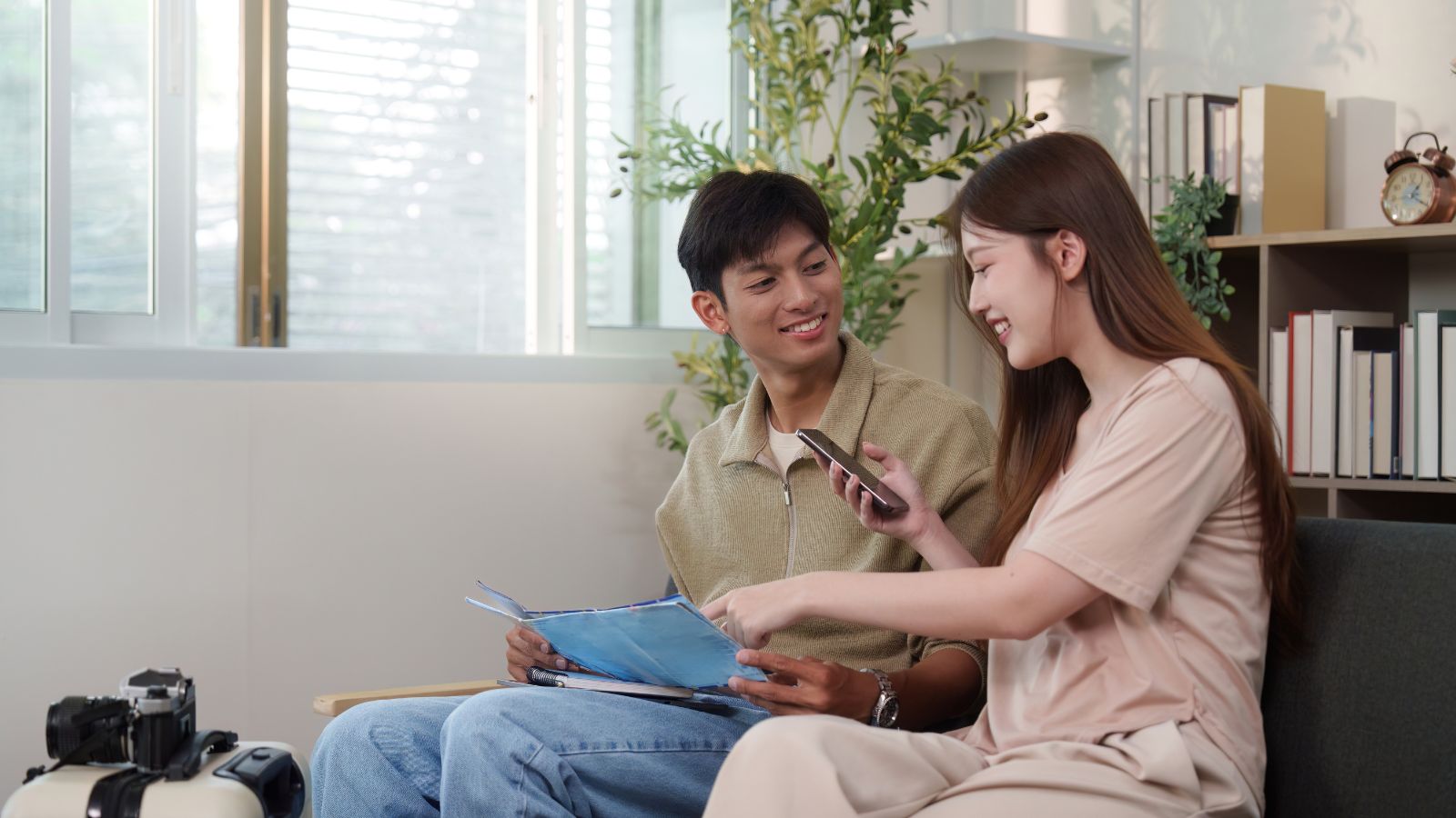 A man with papers and a smiling woman showing her phone sit on a couch, with a camera and books on the nearby table.
