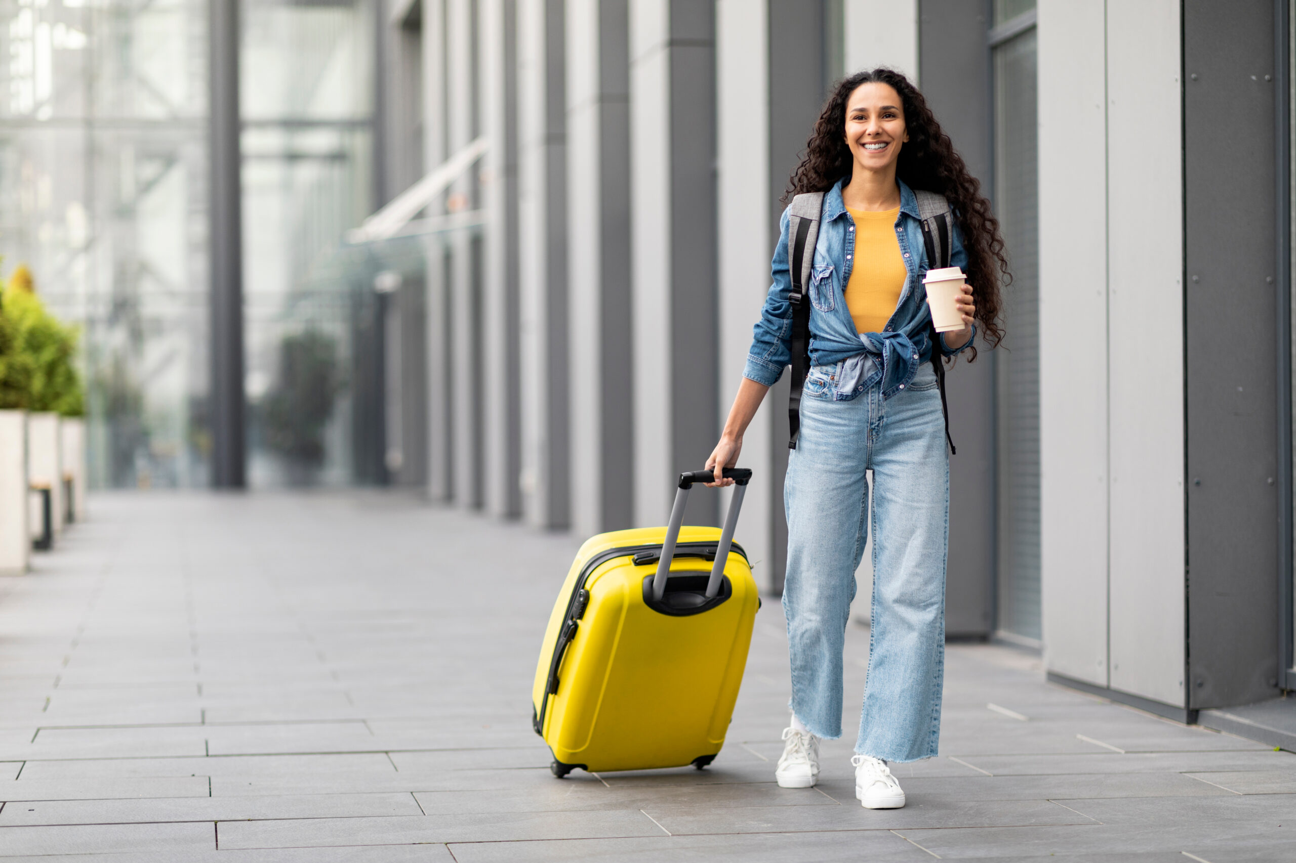 Cheerful pretty long-haired brunette young woman in casual outfit having journey abroad, walking by modern airport with yellow luggage, holding paper cup, drinking coffee, panorama with copy space