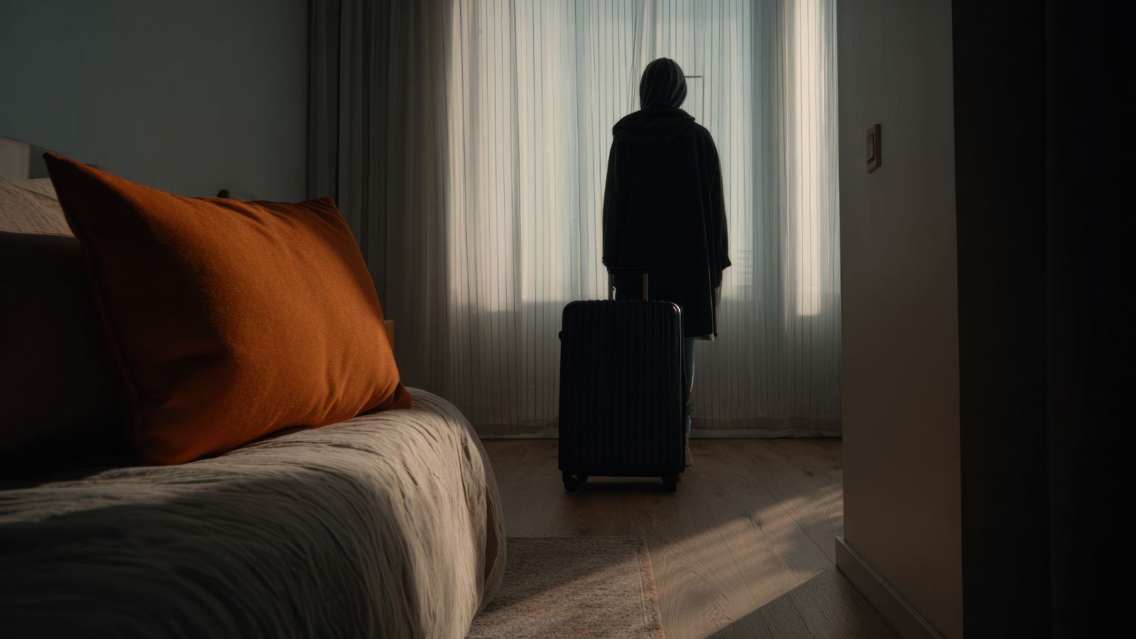 A photo of a traveler, standing with a luggage in her hand, dim setting, facing the curtain.
