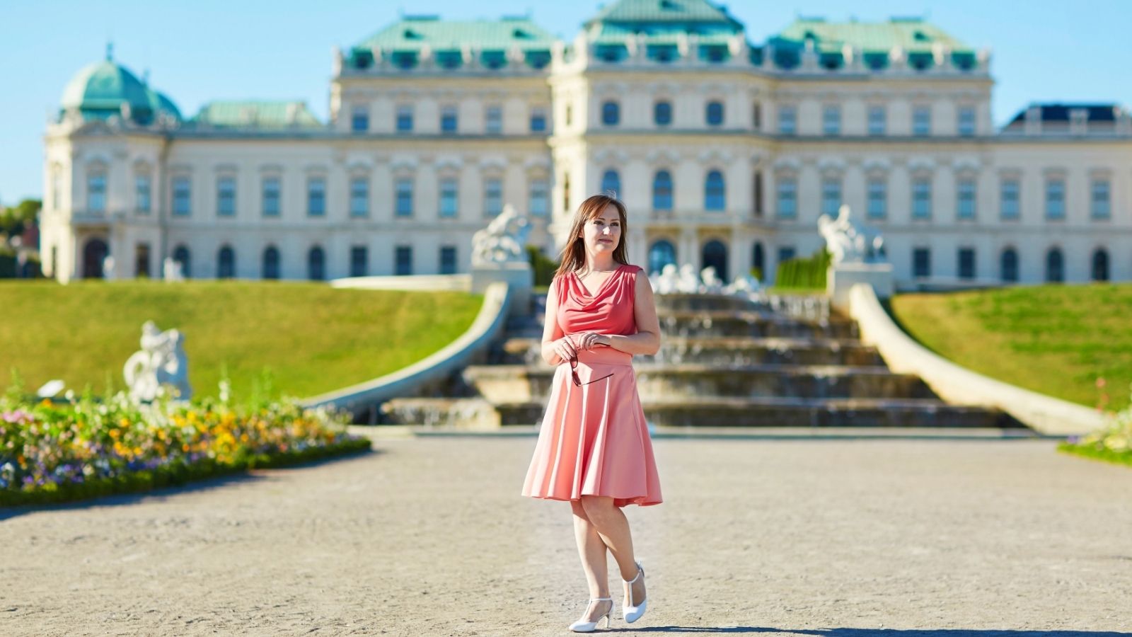 A woman in a pink dress stands before Belvedere Palace with gardens and a fountain visible on a sunny day.