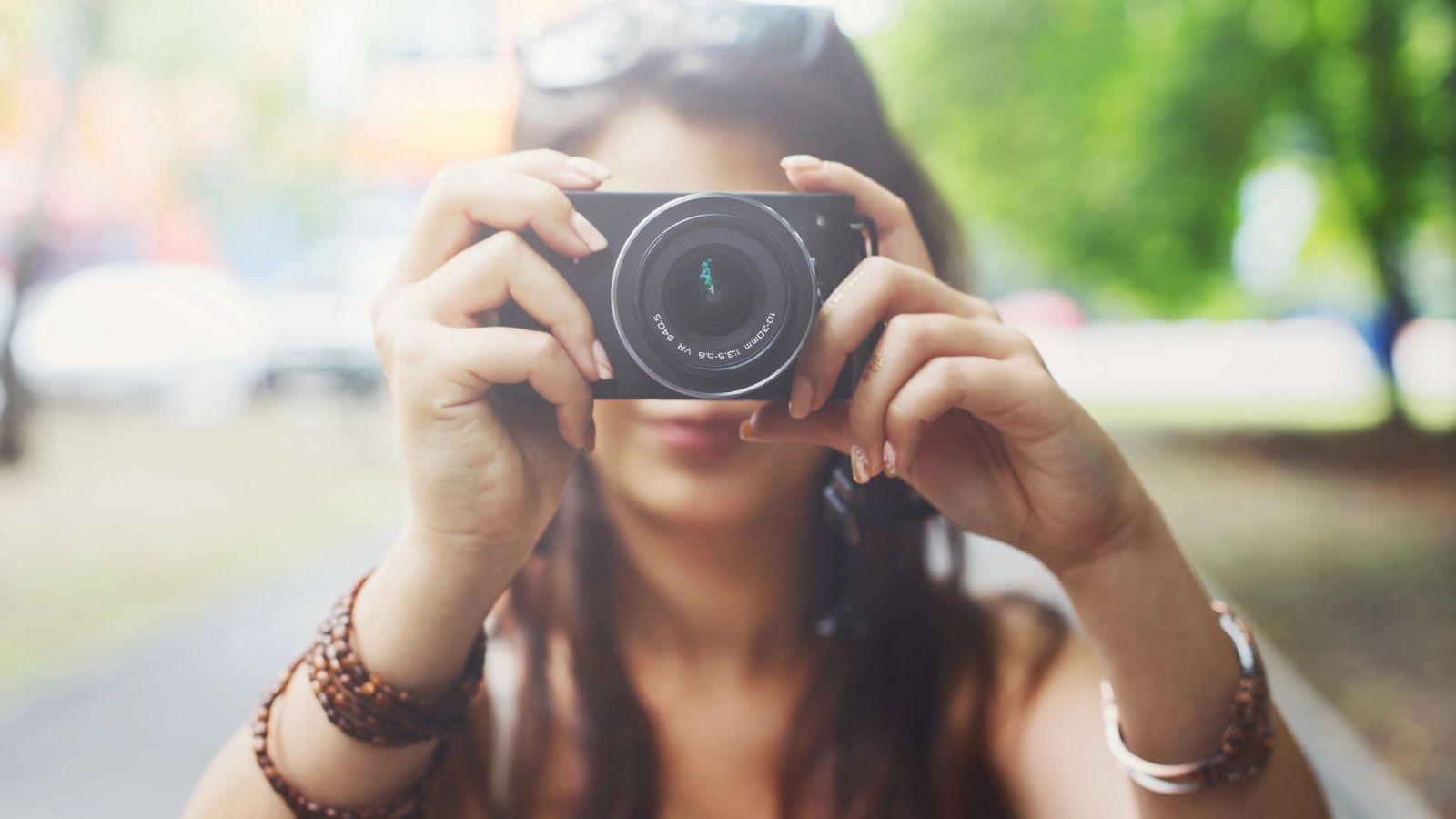 A person outdoors points a camera at the viewer, with blurred greenery and sunlight in the background.
