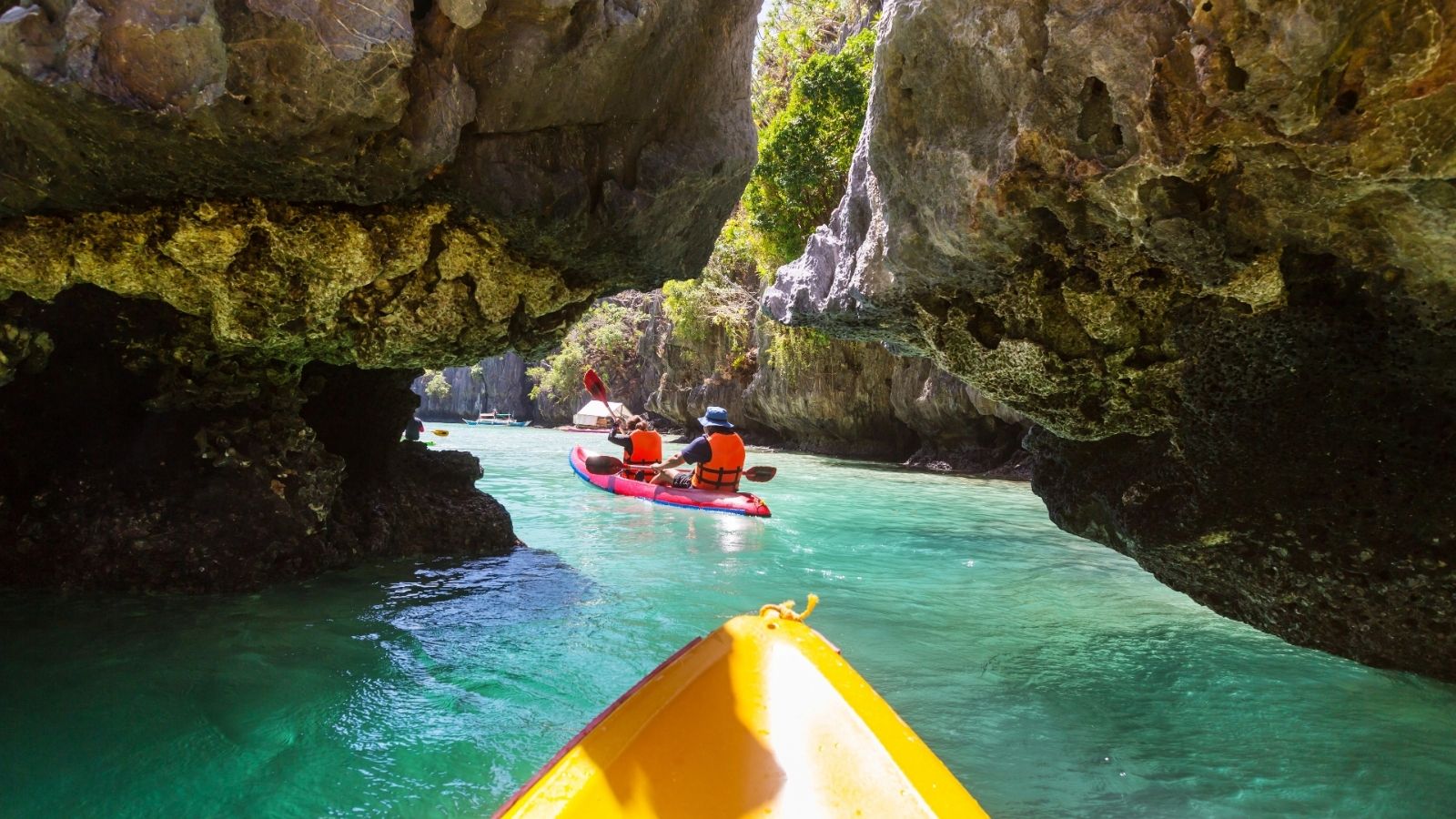 Kayakers glide on clear turquoise water beneath rocky cliffs and caves as sunlight streams through.