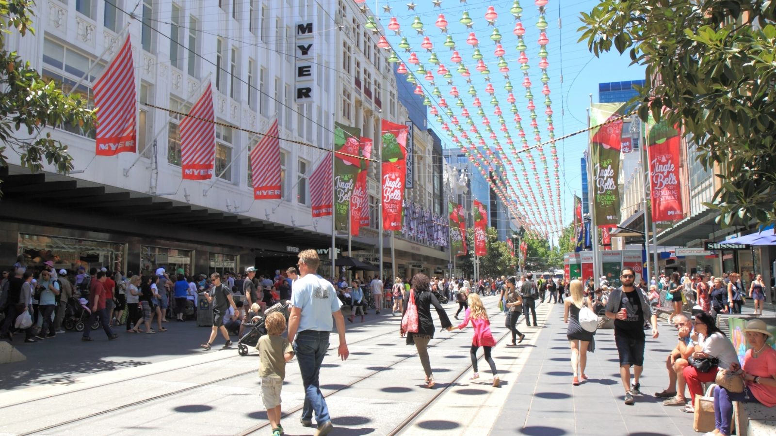 Busy pedestrian street lined with shops and trees, festive banners overhead, people walking beneath a clear sky.