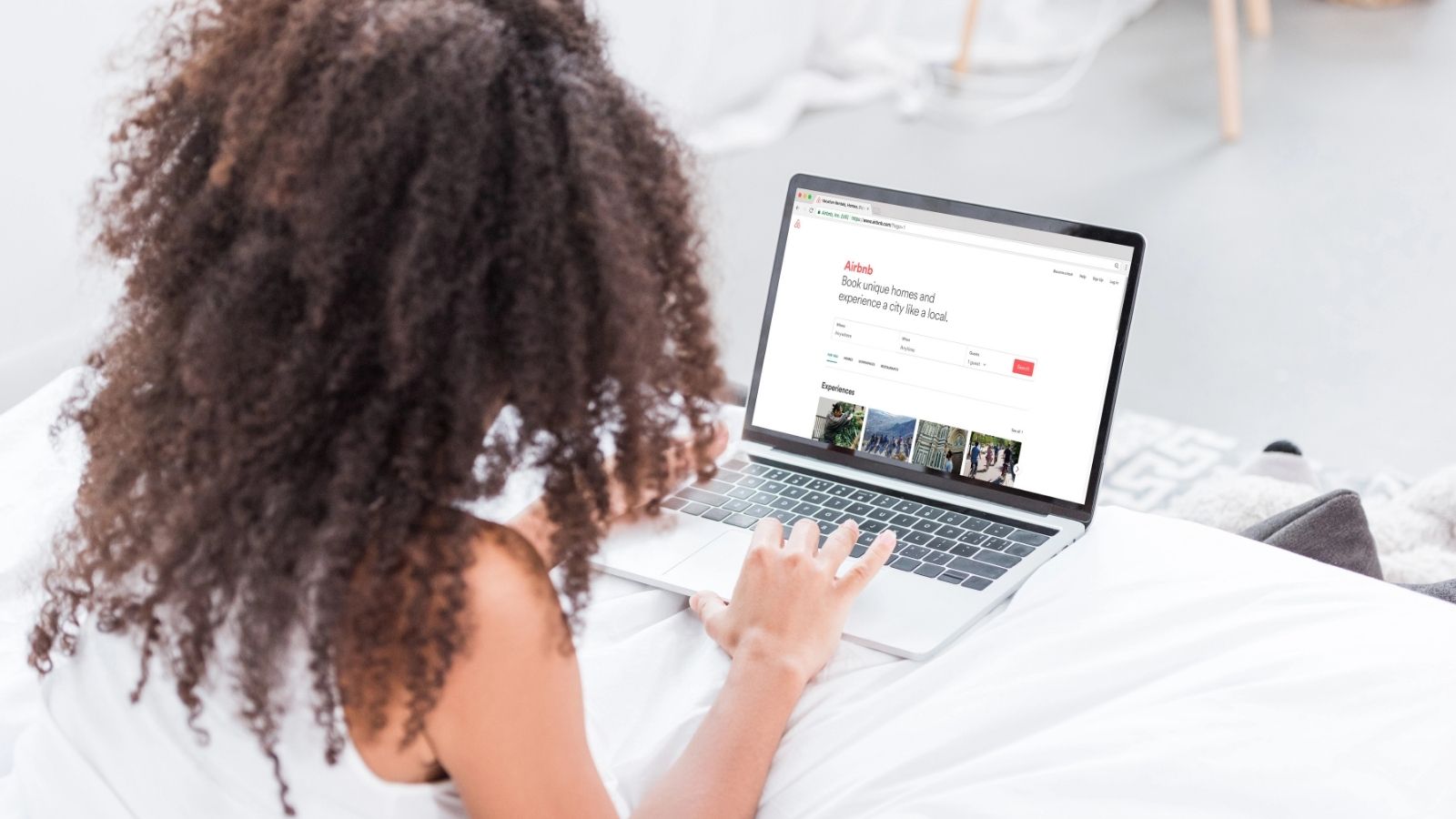 A person with curly hair sits on a bed using a laptop to view the Airbnb website.