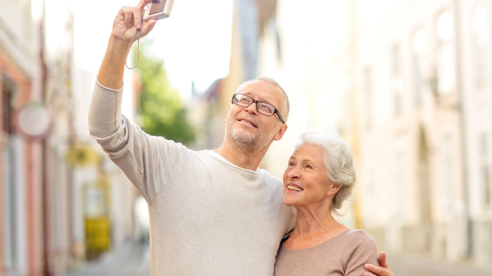An older man and woman smile while taking a selfie together with a digital camera on a city street.