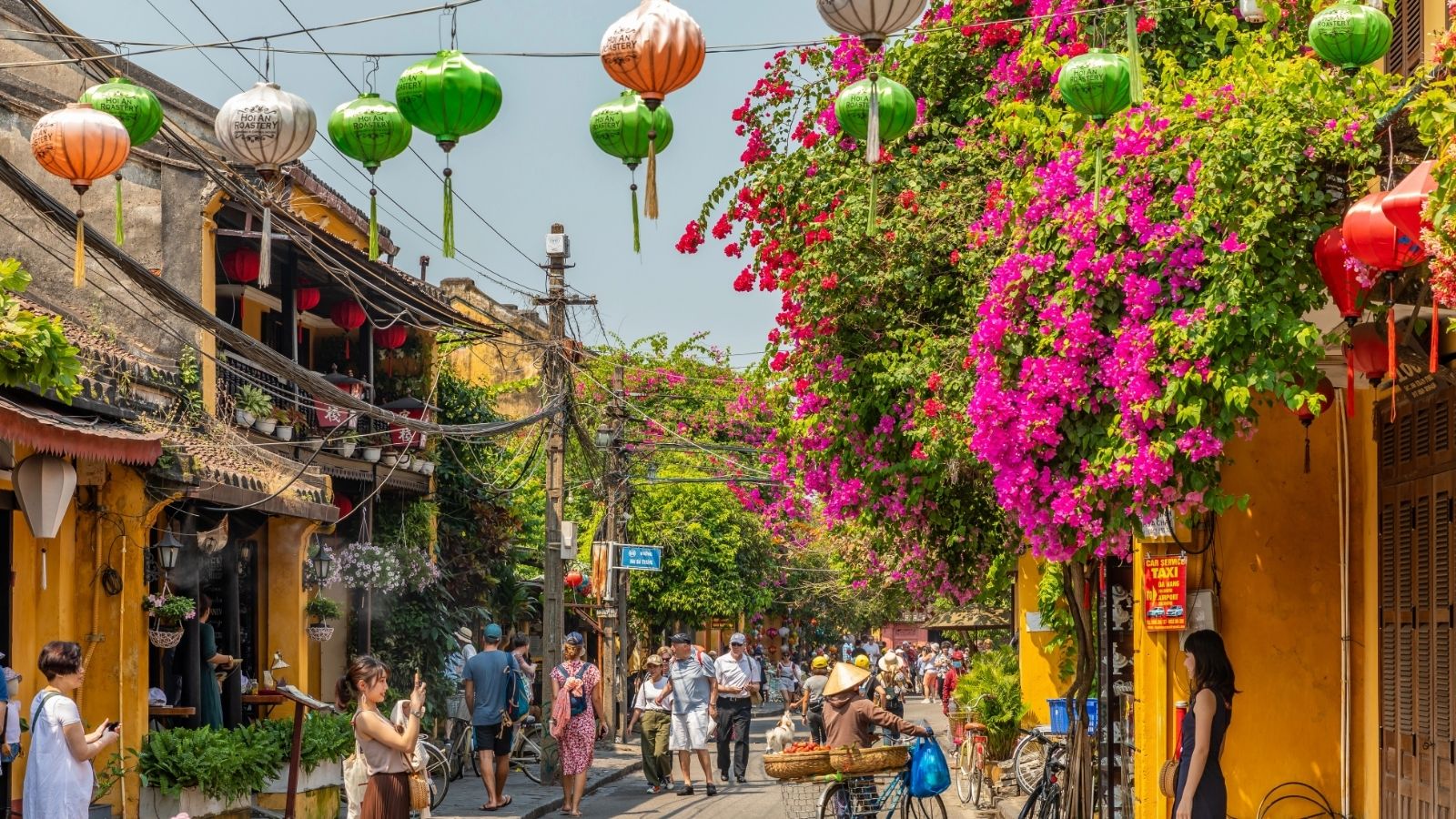 People walk under hanging lanterns beside a building draped in vibrant pink bougainvillea in a lively urban street.