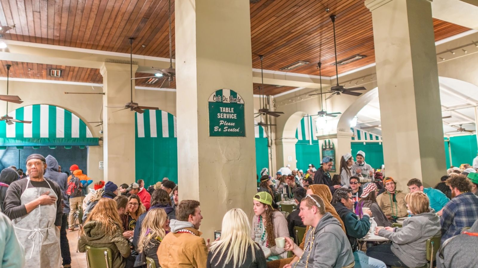 A crowded cafe with diners in jackets and hats, a server on the left, and green-and-white awnings in the background.