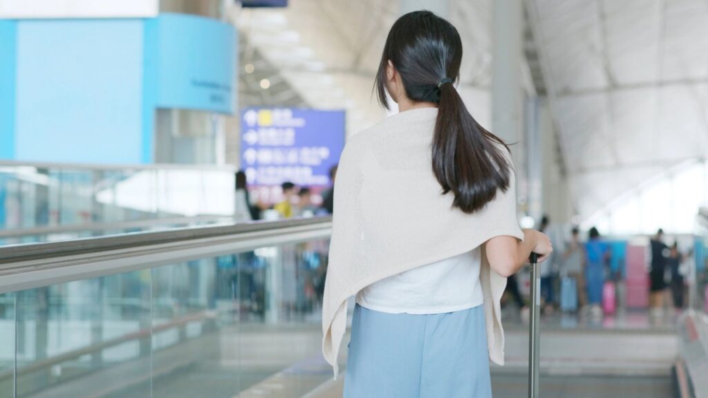 A woman with a ponytail pulls a suitcase on an airport moving walkway, looking ahead at terminal signs.
