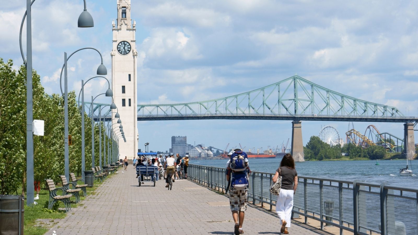 People walk and cycle by a river with benches and trees; a clock tower and steel bridge stand in the partly cloudy background.