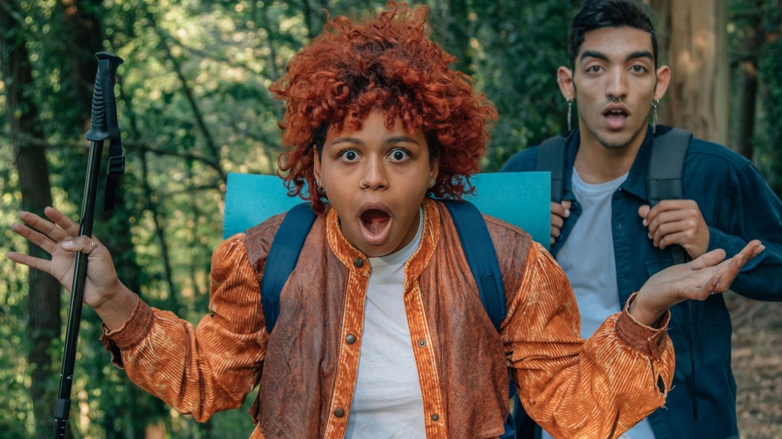 Two hikers in a forest, one with curly red hair and an orange jacket looks surprised as the other stands behind neutrally.