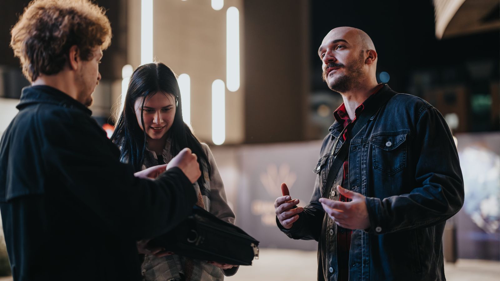 At night, three people stand outside; one opens a briefcase, another looks in, and the third gestures amid blurred city lights.