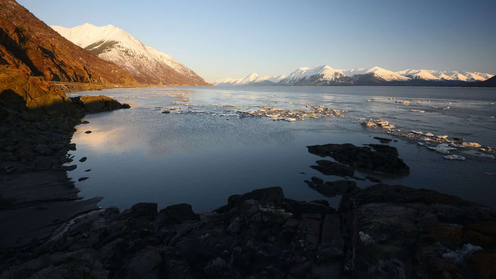 Calm water and rocky shore meet distant snow-capped mountains beneath a clear sky.