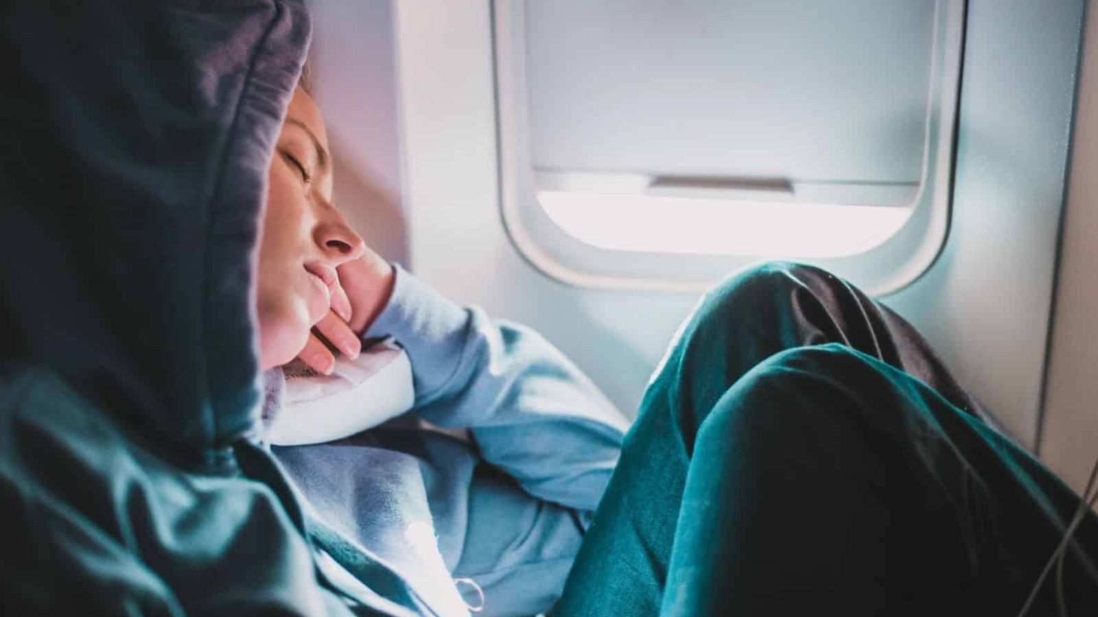 A person in a hoodie sleeps in an airplane seat, head on a pillow by the window and knees pulled to their chest.