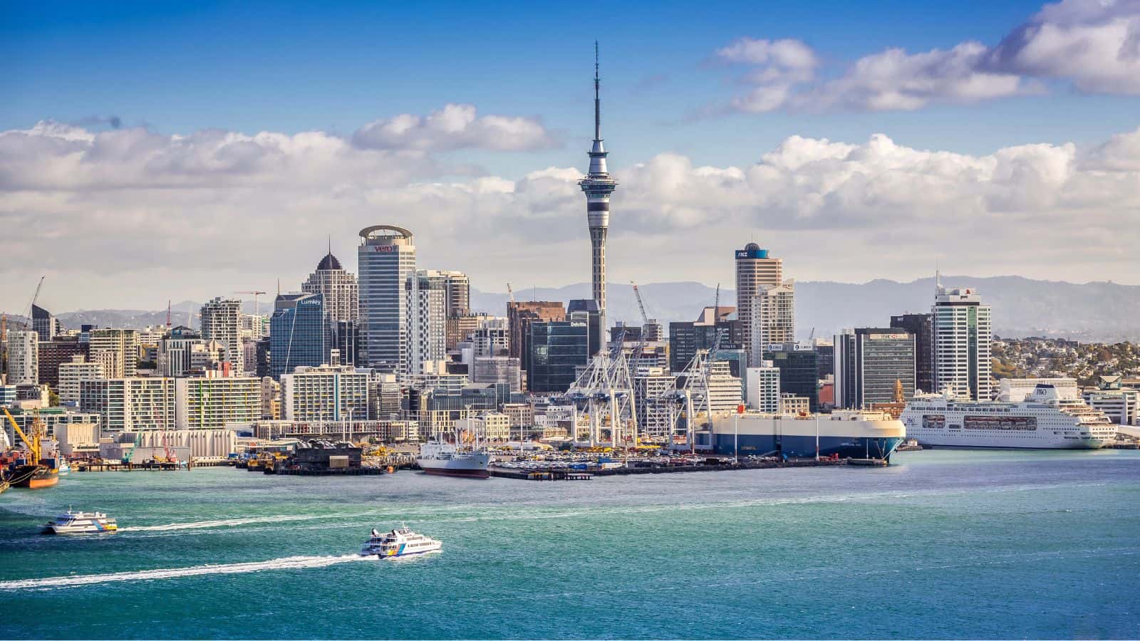 Auckland skyline with Sky Tower, modern buildings, ferries on blue harbor, and cruise ships beneath a partly cloudy sky.