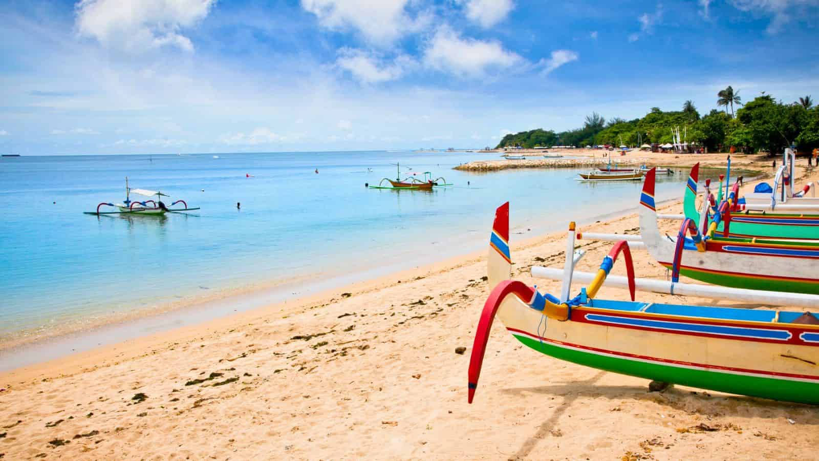 Several colorful wooden boats are lined up on a sandy beach with calm blue waters in the background. A few boats float on the water, and a green, tree-covered shoreline is visible under a bright, partly cloudy sky.
