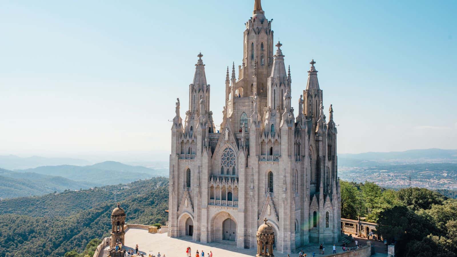 A large, ornate church with Gothic-style architecture stands atop a hill, surrounded by greenery and distant mountains under a clear blue sky. A few people walk near the entrance.