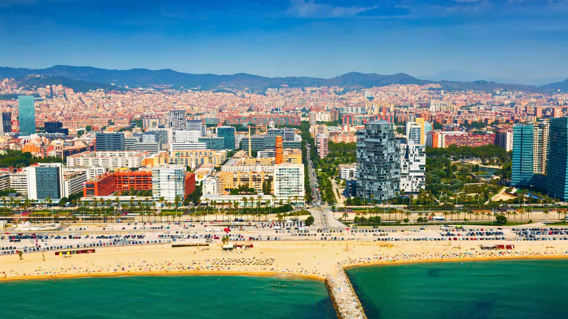 Aerial view of a city with modern buildings, a sandy beach, and clear blue-green sea under a sunny sky.