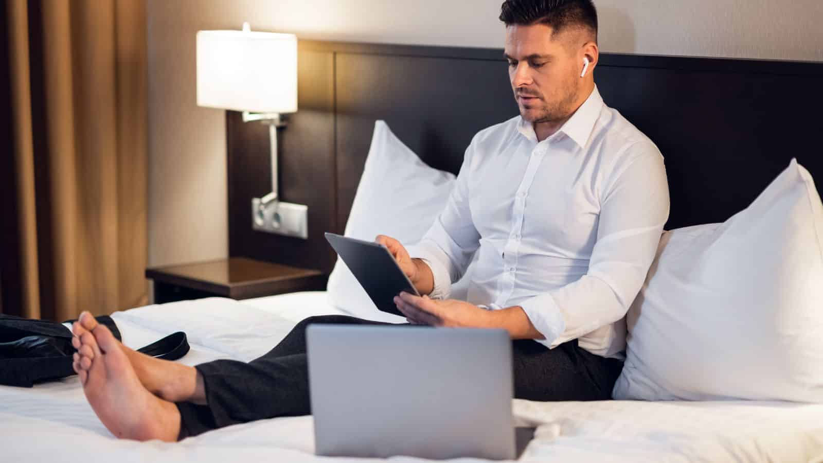 A man in business attire sits on a hotel bed, focused on his laptop and tablet while wearing wireless earbuds.