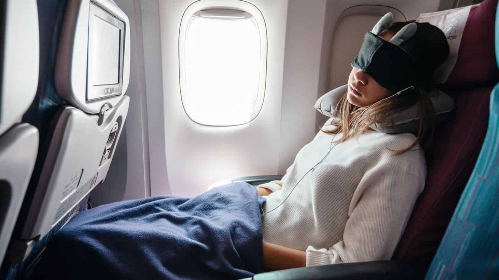 A person sleeps in an airplane seat by the window, wearing an eye mask, headphones, neck pillow, and covered with a blue blanket.